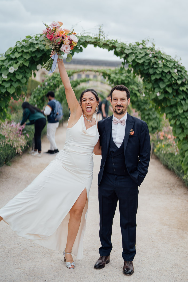 Une mariée exaltée levant son bouquet avec enthousiasme, accompagnée de son époux souriant.