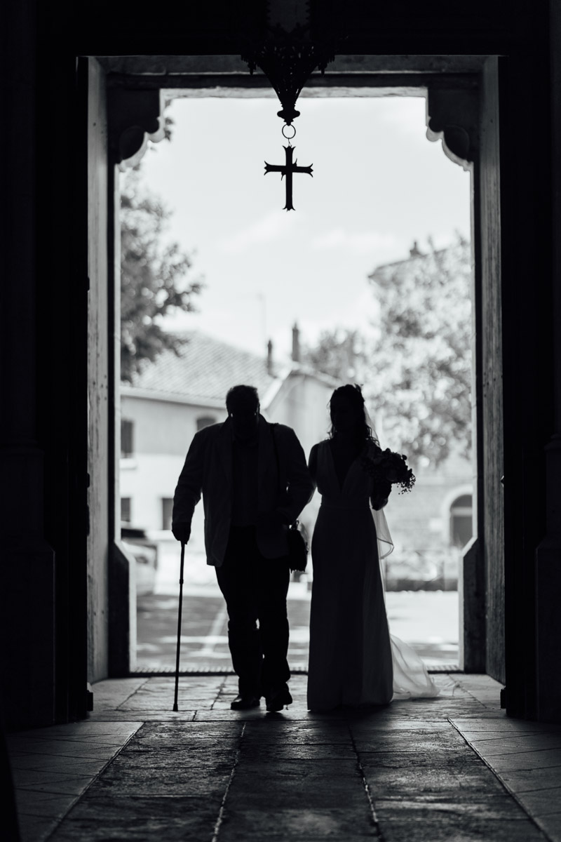 Une mariée entrant dans l’église au bras de son père dans un jeu de lumière en contre-jour.