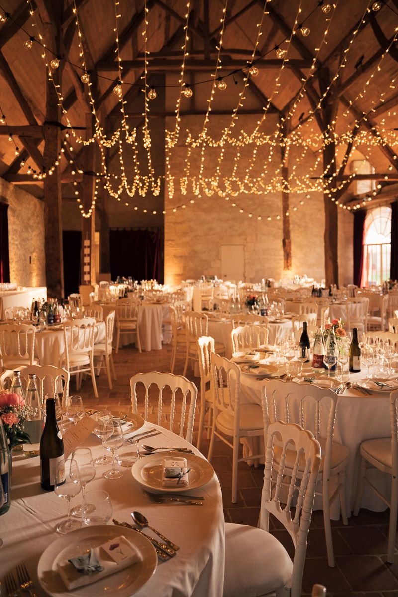 Salle de réception rustique éclairée par des guirlandes lumineuses avec tables dressées pour le mariage