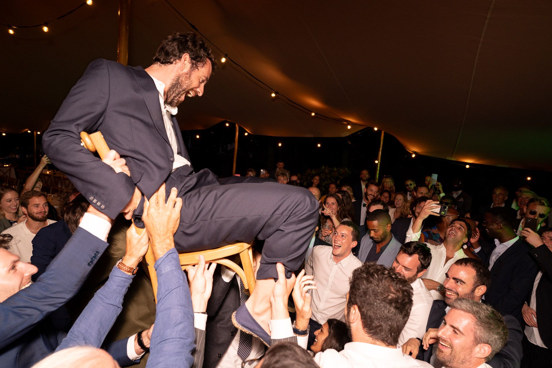 Photo d'une soirée de mariage festive et animée. Le marié est porté sur une chaise au-dessus de ses invités.