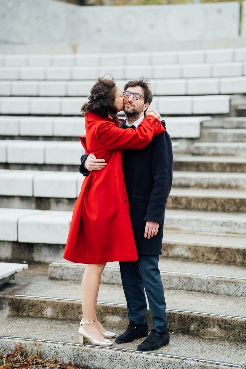 Couple en manteaux sur des marches, femme en rouge embrassant homme à lunettes.